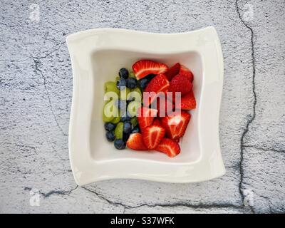 Nahaufnahme von frischen Sommerbeeren in einer weißen Schüssel vor einem strukturierten natürlichen gerissenen Steinhintergrund. Erdbeeren, Heidelbeeren und Trauben auf der Küchentheke zubereiten Stockfoto