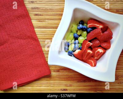 Nahaufnahme von frischen Sommerbeeren in einer weißen Schüssel gegen ein rustikales Holzbrett. Erdbeeren, Heidelbeeren und Trauben auf der Küchentheke zubereiten. Stockfoto