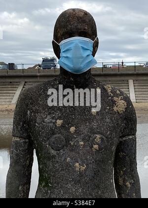 Antony Gormley’s Another Place am Crosby Beach, Liverpool, wo eine der lebensgroßen Metallstatuen mit einer Gesichtsmaske und einer Referenz auf das Coronavirus geschmückt wurde. Stockfoto