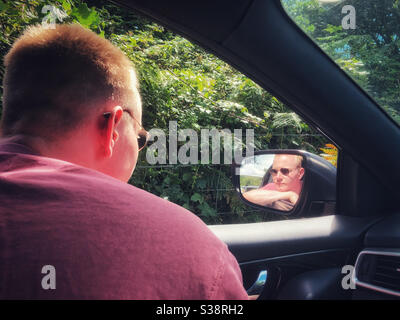 Junger Mann, der aus einem Autofenster schaute und sich im Außenspiegel spiegelte. Stockfoto