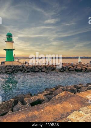 Der grüne Leuchtturm am Wellenbrecher der deutschen Ostseestadt Warnemünde im Abendlicht mit Segelbooten, Menschen und einem roten Turm im Hintergrund Stockfoto