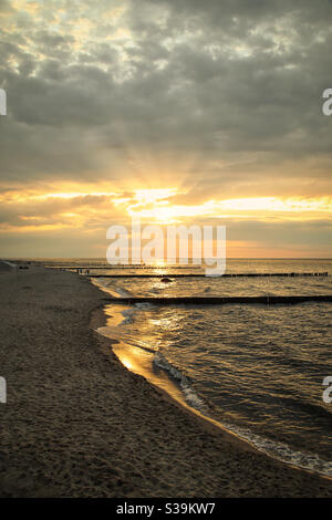 Letzte Sonnenstrahlen an einem Ostseestrand. Im Hintergrund sieht man Spaziergänger und Liegestühle und die typischen Groynes am Ufer Stockfoto