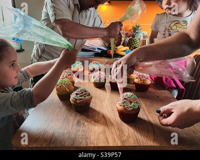 Gruppe von Kindern backen Cupcakes, Quetschen Sahne aus Süßwarenbeutel, Zutaten vorbereiten, Belag, Streuseln für die Dekoration von Cookies. Kinder kochen, arbeiten zusammen in der Küche zu Hause Stockfoto