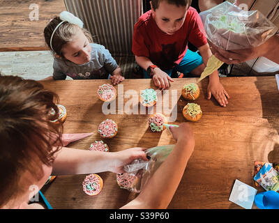 Gruppe von Kindern backen Cupcakes, Quetschen Sahne aus Süßwarenbeutel, Zutaten vorbereiten, Belag, Streuseln für die Dekoration von Cookies. Kinder kochen, arbeiten zusammen in der Küche zu Hause Stockfoto