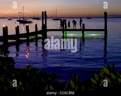 Silhouette von mehreren Menschen, die nach Sonnenuntergang auf einem Pier mit Segelbooten im Wasser stehen. Stockfoto