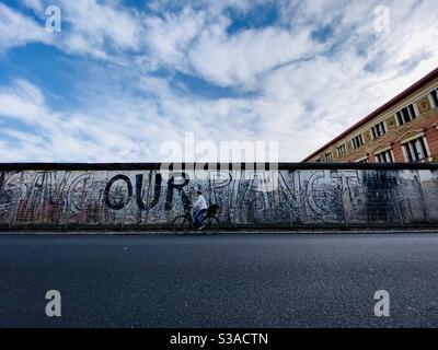 Eine Frau fährt mit dem Fahrrad entlang eines Teils der Berliner Mauer, der noch oben ist. Graffiti an der Wand lautet „rette unseren Planeten“. Berlin, Deutschland. Stockfoto