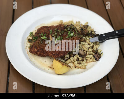 Gebratenes Hühnchen-Schnitzel mit Zitrone und Spatzle-Pasta mit Pilzen werden auf einem Teller in einem Restaurant gezeigt. Diese aktualisierte Version des deutschen Klassikers hat Kapern und und Schalotten auf dem Huhn. Stockfoto