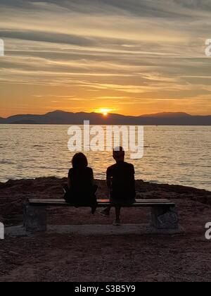 Zwei Personen sitzen zusammen auf einer Bank, umgeben von einem atemberaubenden Sonnenuntergang über dem Meer, mit fernen Bergen, die eine ruhige Landschaft schaffen Stockfoto