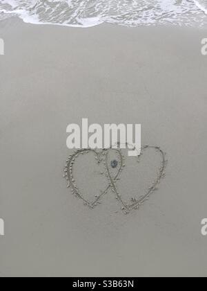Herzen im Sand, aber nicht lange. Eingehende Kurven werden überspült und die Zeichnung in kurzer Reihenfolge gelöscht. Diese Kunst ist nicht lang und die Zeit vergeht. Stockfoto