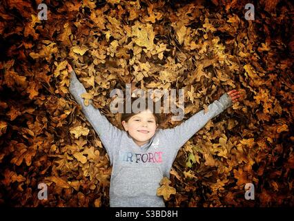 Kleines Mädchen, das an einem Herbsttag fröhlich in einem riesigen Laubhaufen spielt. Stockfoto