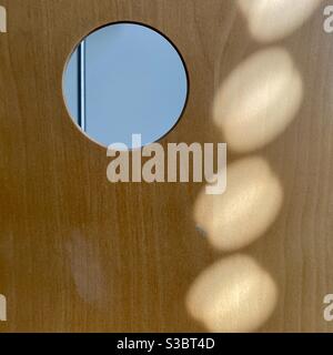 Lichtmuster, das durch Sonneneinstrahlung auf die Rückseite eines gebogenen Holzstücks mit einem Loch oben rechts und einem unscharf dahinter liegenden Objekt entsteht, was einen interessanten abstrakten Hintergrund erzeugt Stockfoto