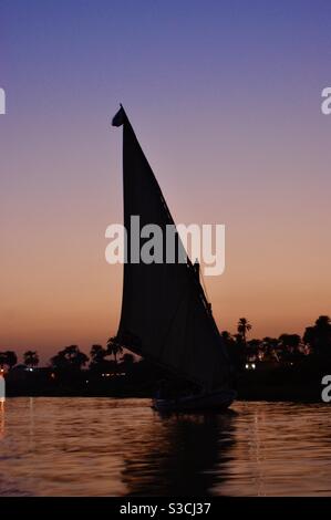 Silhouette eines traditionellen Felucca-Flußbootes, das während eines spektakulären Sommeruntergangs entlang des Nils segelt und den Himmel in einem weichen orange, rosa, pfirsichfarbenen und violetten Schein in Luxor, Ägypten, beleuchtet. Stockfoto