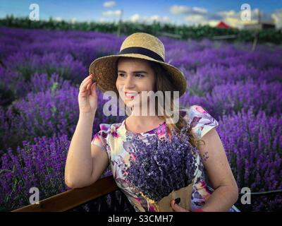 Porträt einer glücklichen Frau mit Strohhut mit einem Strauß Von Lavendel, während Sie sich auf einer Bank in einem Lavendelfeld Stockfoto