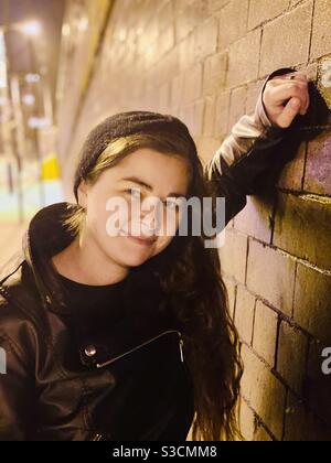 Eine junge Frau lehnt sich an eine Backsteinmauer mit gelben Lichtern, die hinter ihr hereinströmen. Stockfoto