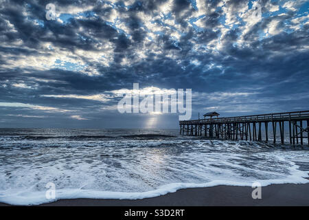 Crepuscular Strahlen scheinen durch die Wolken über dem Amelia Island Beach Pier mit Wellen Rollen in. Stockfoto