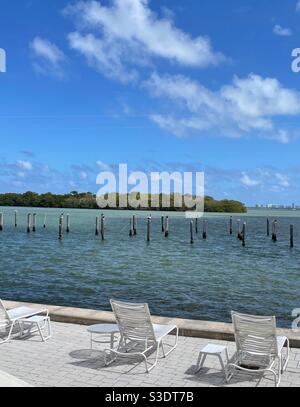 Weiße, flauschige Wolken in einem blauen Himmel werfen Schatten auf das türkisfarbene Meerwasser von Miami Beach, Florida, USA in einer ikonischen Umgebung mit weißen Liegestühlen mit Blick auf eine tropische Insel und alte Pier Pilings. Stockfoto