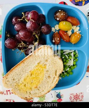 Ein einfaches, aber gesundes Mittagessen für ein Kind mit frischen roten Trauben, Kirschtomaten mit Feta-Käse, frischen Erbsen mit Mozzarella und einer Scheibe Toast mit Butter auf einem blauen Teller Stockfoto