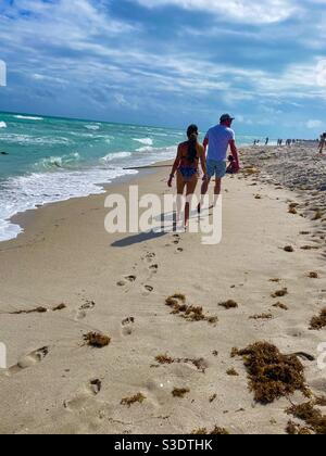 Ein Mann und eine Frau hinterlassen Fußspuren im Sand, während sie am Strand neben weißgedeckten Wellen auf dem türkisfarbenen Meer in Miami South Beach, Florida, USA, spazieren Stockfoto