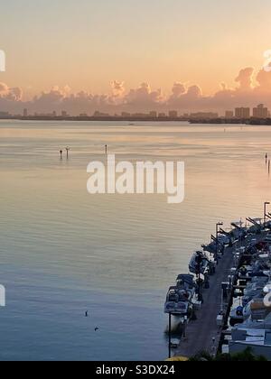 Goldrosa Sonnenaufgang mit wabenden Wolken am Horizont über stillem Meereswasser mit geparkten Booten im Vordergrund und Hochhäusern im Hintergrund in Miami Beach, Florida, USA Stockfoto