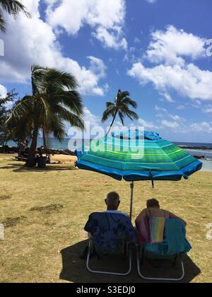 Ein sonniger Tag am Strand, Maui, Hawaii. Ein Paar bleibt unter dem Sonnenschirm kühl. Sonnenanbeter auf dem Wasser. Winterzeit. Die Weihnachtszeit. Stockfoto