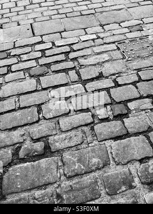 Kopfsteinpflasterstraße (und Wasserschacht) in Wells, Somerset, Großbritannien, Kopfsteinpflaster, Straße, Bürgersteig, holprig, uneben, alt Stockfoto