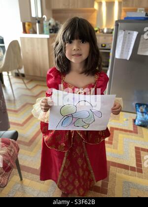 Schöne und nette gemischte asiatische kleine Mädchen mit langen braunen Haaren trägt ein rotes Kleid mit goldenen Zierleisten hält eine schöne Farbzeichnung, lächelnd Stockfoto