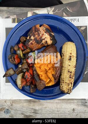 Eine köstliche Mahlzeit aus Lachs, Maiskolben, Süßkartoffeln und gemischtem Gemüse, gegrillt am offenen Feuer, serviert auf einem blau emaillierten Blechteller. Stockfoto