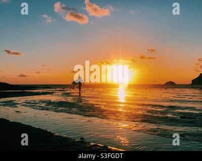 Ein Surfer spaziert am Strand entlang, vor einem wunderschönen Sonnenuntergang in Cornwall, England Stockfoto