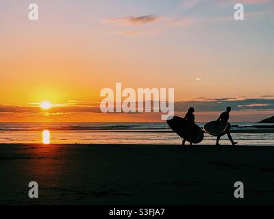 Surfer, die am Strand in Devon, Großbritannien, spazieren, vom Sonnenuntergang geschildet Stockfoto