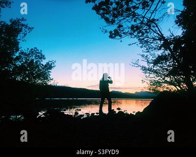 Mann trinkt ein Bier und beobachtet den Sonnenuntergang über dem Lake Windermere Stockfoto