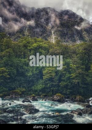 Neblige Berge, Wasserfälle und blauer Alpenbach im Fiordland National Park, Südinsel, Neuseeland Stockfoto