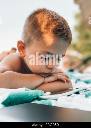 Der kleine fünfjährige Junge lag am Pool und beobachtete einen Cartoon am Telefon Stockfoto