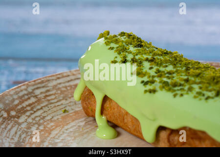 Pistazie eclair bestreut mit grünen Spanplatten auf einem Teller mit blauem Hintergrund. Süßwaren. Stockfoto