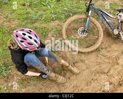 Blick aus der Perspektive auf ein Mädchen, das mit Schlamm bedeckt ist und auf dem Boden neben ihrem schlammigen Mountainbike sitzt Stockfoto