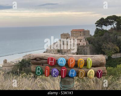 Kreatives Happy Birthday mit farbigen Steinbuchstaben vor einem mittelalterlichen Dorf mit Meereshintergrund Stockfoto
