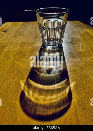 Ein Glas Wasser steht auf einem Holztisch und wirft einen langen Schatten Stockfoto