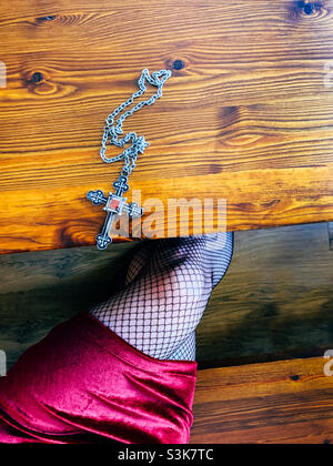 Halskette mit silbernem Kreuz auf einem Holztisch neben einer Frau, die ein rotes Samtkleid und Netzstrumpfhosen trägt Stockfoto