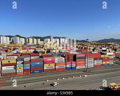 Busan New Port, Südkorea Blick von der Brücke aus vom festfahrenden Handelsfrachtschiff auf die im Containerterminal verstauten Container. Im Hintergrund befinden sich Häuser und Wohnblocks von Hafenarbeitern. Stockfoto