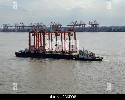 Schlepper auf dem Yangtze-Fluss in der Nähe des Hafens von Shanghai, der das Schiff mit neuen Straddle-Trägern beladen hat, die für den Transport von Containern in Terminals verwendet werden. Im Hintergrund befinden sich Portalkrane an der Küstenlinie. Stockfoto