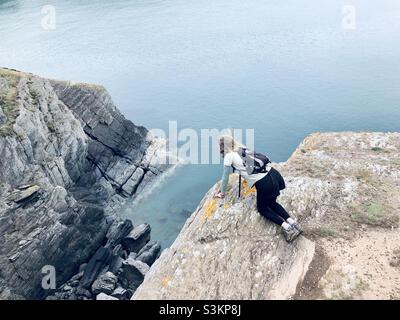 Eine Frau in einem kleinen Rucksack blickt über den Rand einer Klippe am Meer in Pembrokeshire in Wales Stockfoto