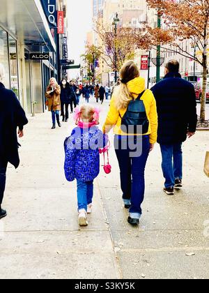 Ein süßes kleines Mädchen mit rosa Bändern im Haar geht mit anderen Fußgängern auf einem Bürgersteig in New York City Stockfoto