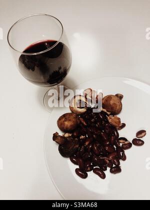 Ein Glas Rotwein und einige Kidney-Bohnen und Pilze auf einem weißen Teller auf weißem Hintergrund serviert Stockfoto