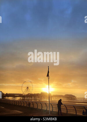 Figur in Silhouette, die an einer Barriere angelehnt ist und die Nachmittagssonne hinter dem Central Pier in Blackpool beobachtet Stockfoto