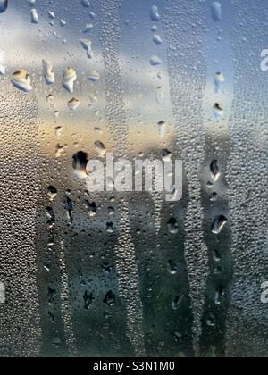 Wassertröpfchen und Rinnen auf einer mit Kondenswasser bedeckten Fensterscheibe vor einem Morgenhimmel Stockfoto