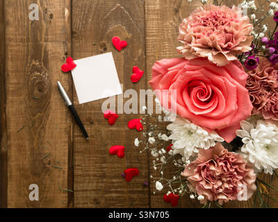 Draufsicht auf einen Holztisch, auf dem sich ein Strauß in einer Vase mit verschiedenen Blumen befindet, ein kleines Stück Papier für Notizen und ein Kugelschreiber liegen in der Nähe, rote Herzen sind auf der Oberfläche verstreut. Stockfoto