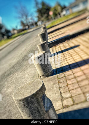 Ein geneigter Blick an einem sonnigen Tag auf eine Reihe von runden Betonbarrieren, die einen öffentlichen Fußweg von einer Straße trennen, wobei sich lange, enge Schatten von jedem von ihnen erstrecken. Stockfoto
