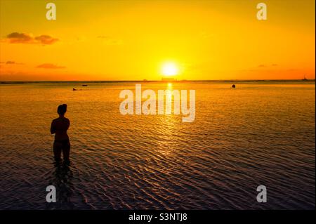 Eine Dame, die im Meer steht und den Sonnenuntergang vor dem Hintergrund der Silhouette des Meeres im Ala Moana Beach Park beobachtet Stockfoto