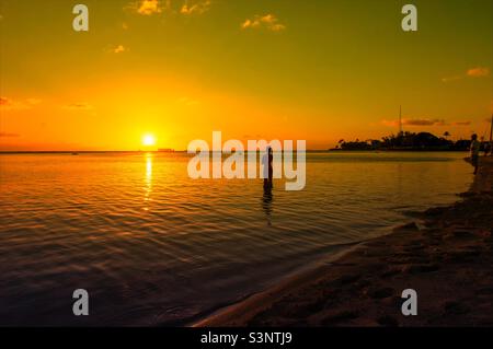 Eine Dame, die im Meer steht und einen wunderschönen Sonnenuntergang mit Sonnenuntergangshintergrund im Ala Moana Beach Park in Honolulu City Oahu Island Hawaii beobachtet Stockfoto