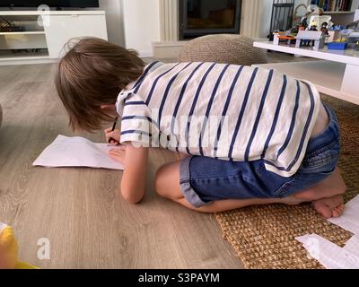 Kleiner Junge kniet auf dem Boden und zeichnet auf weißem Papier Stockfoto