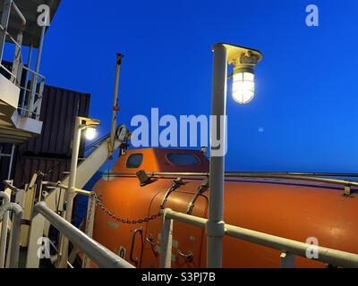 Orange Rettungsboot in Hafenseite des Containerschiffes befestigt bereit für den Notfall beleuchtet von zwei elektrischen Lampen während des Sonnenuntergangs . Das Schiff segelt im Mittelmeer bei ruhigem Frühlingswetter. Stockfoto